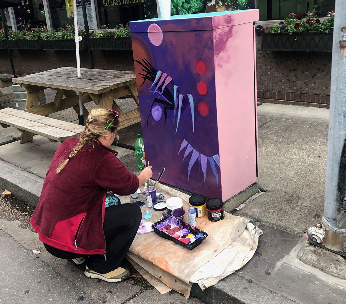 woman painting electrical box with purple and reds.